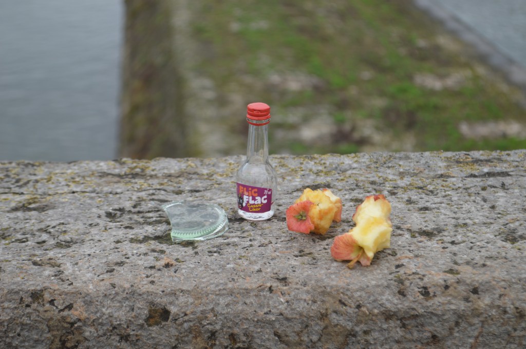 Found Art in Würzburg, 2019 Photograph In Würzburg, there is a bridge where tourists can drink wine purchased at nearby stores. I found this assortment of trash and found it humorous.